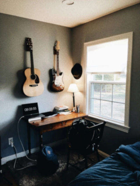 A cozy student room with guitars mounted on the wall, a wooden desk with a laptop, and a window letting in natural light.