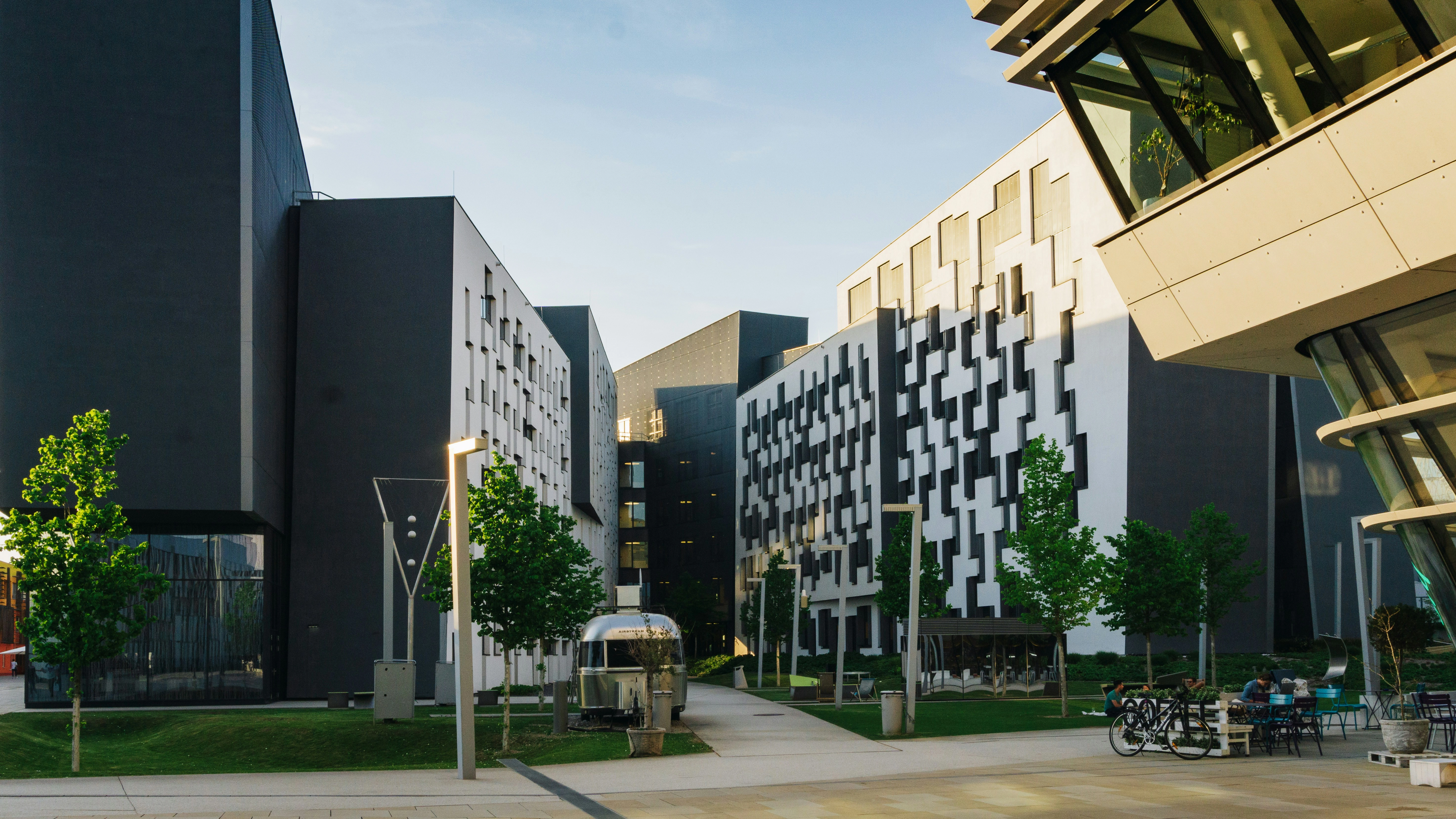 Modern student housing complex with geometric black and white building facades, featuring a green courtyard with trees, outdoor seating, and bicycles parked in the foreground.